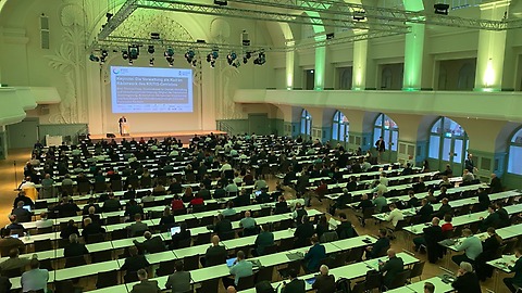 Ein großer Hörsaal mit vielen langen Tischreihen ist mit zahlreichen Personen besetzt, die einer Präsentation auf einer Bühne mit projizierter Folie folgen; der Saal ist hoch, weiß gestaltet und grün beleuchtet. Ein großer Hörsaal mit vielen langen Tischreihen ist mit zahlreichen Personen besetzt, die einer Präsentation auf einer Bühne mit projizierter Folie folgen; der Saal ist hoch, weiß gestaltet und grün beleuchtet.